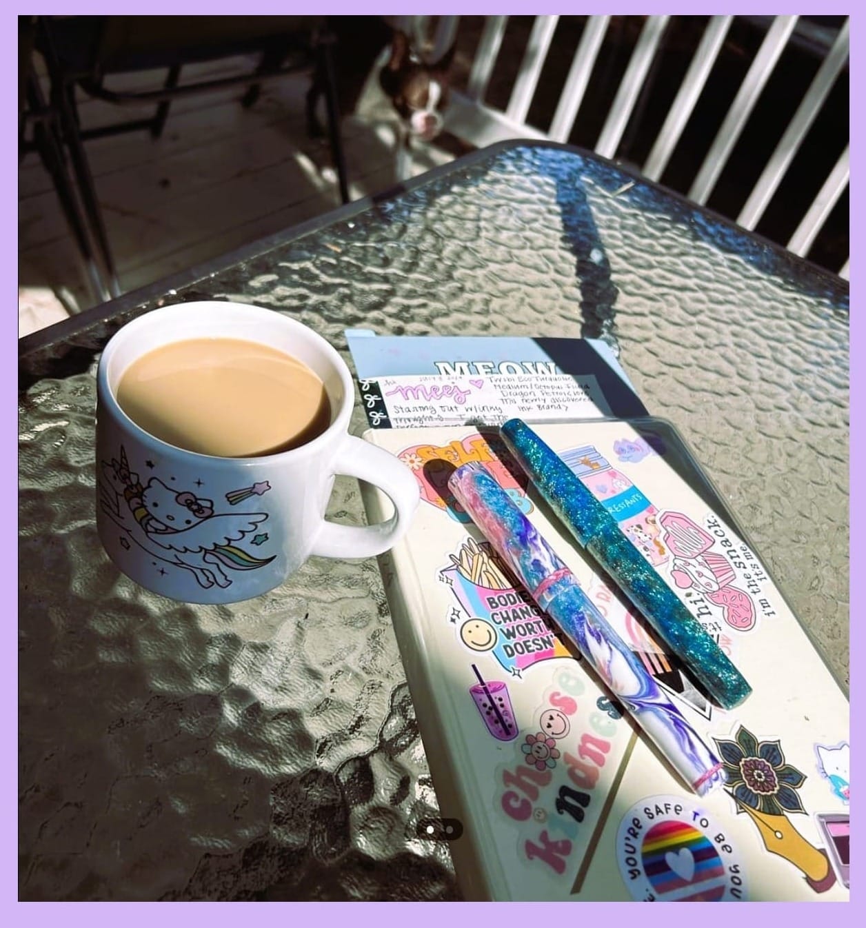 Photo of a glass table with a cup of coffee & a closed planner with various stickers on the front. On top of the planner are 2 fountain pens. There is an out-of-focus dog at the top.